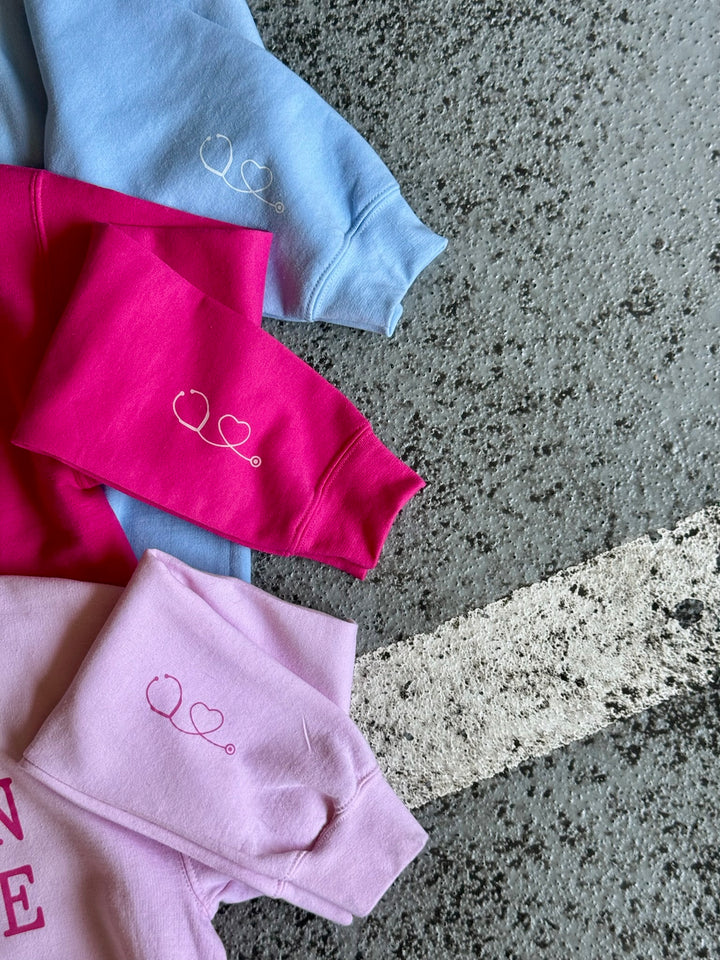 Close up view of the sleeves of the Light Pink, Light Blue, and Hot Pink Women in Medicine Crewneck Sweatshirts showing the Stethoscope Design.