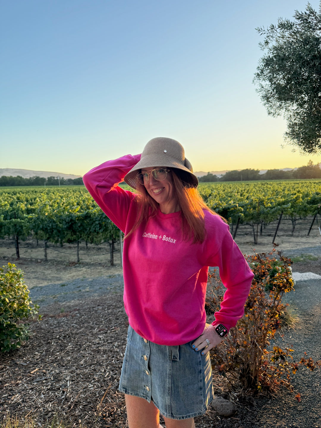 Ladyspinedoc wearing a Dr. Daisy Collab pink Caffeine + Botox sweatshirt and denim skirt standing in a vineyard.