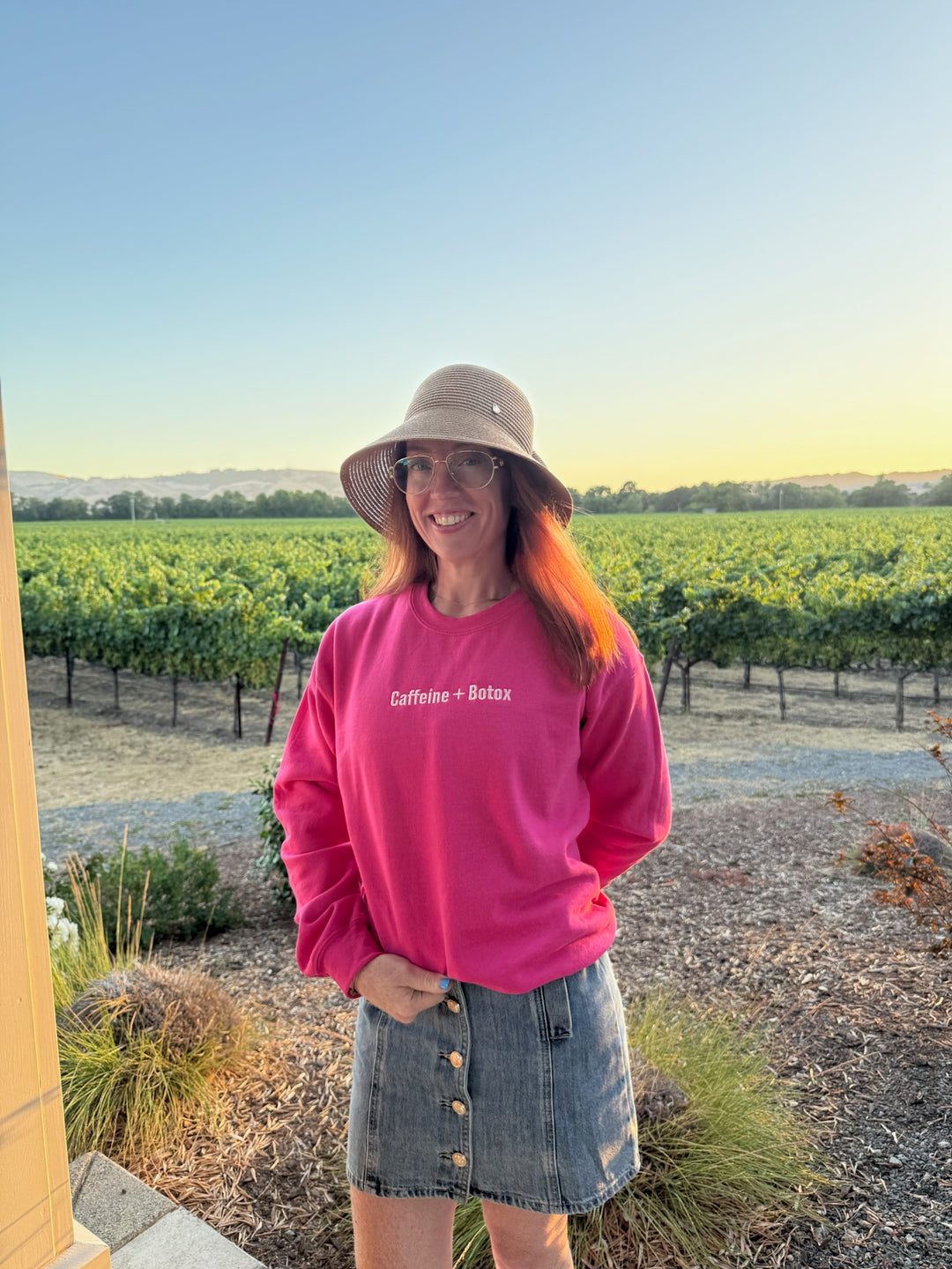 Ladyspinedoc wearing a pink Caffeine + Botox sweatshirt and sun hat in a vineyard setting