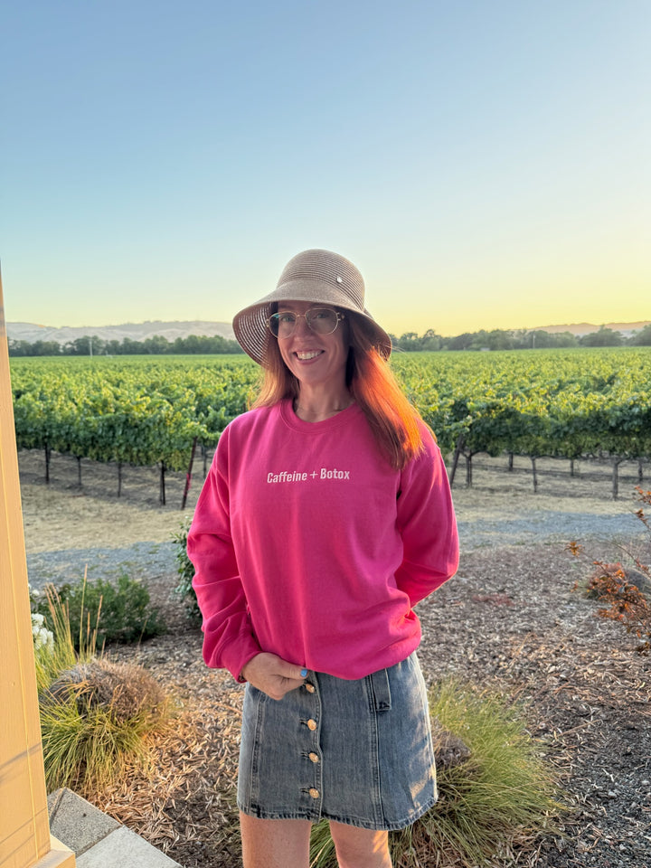 Ladyspinedoc wearing a pink Caffeine + Botox sweatshirt and sun hat in a vineyard setting