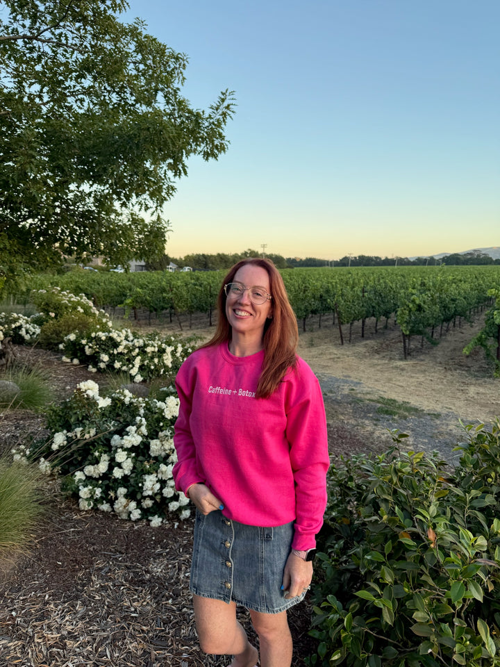 Ladyspinedoc wearing a pink Caffeine + Botox sweatshirt standing in a vineyard with greenery and flowers around.