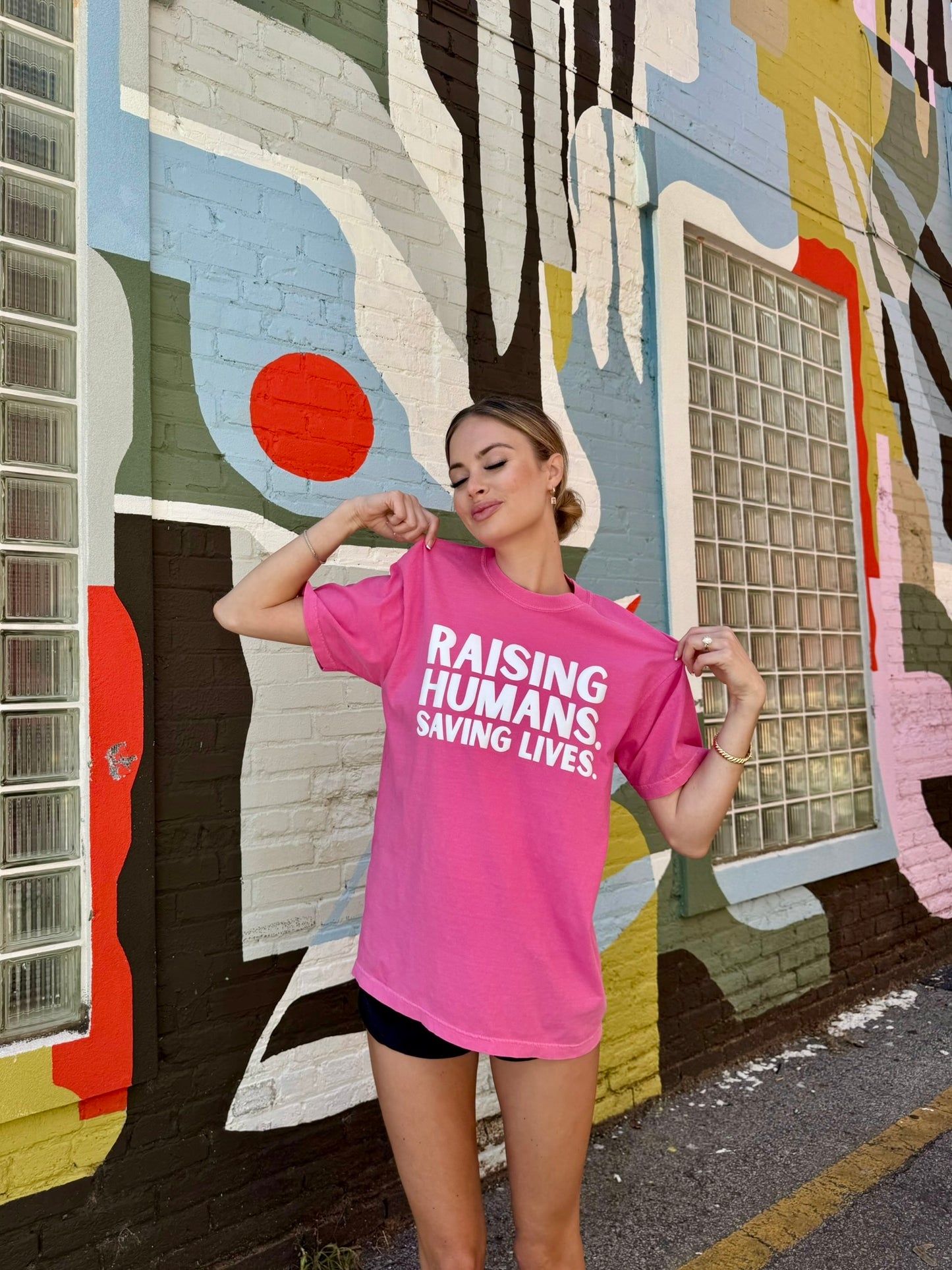 Woman wearing pink Raising Humans. Saving Lives. tee by Ladyspinedoc, holding the shoulders of the shirt with a confident smile in front of a vibrant mural.