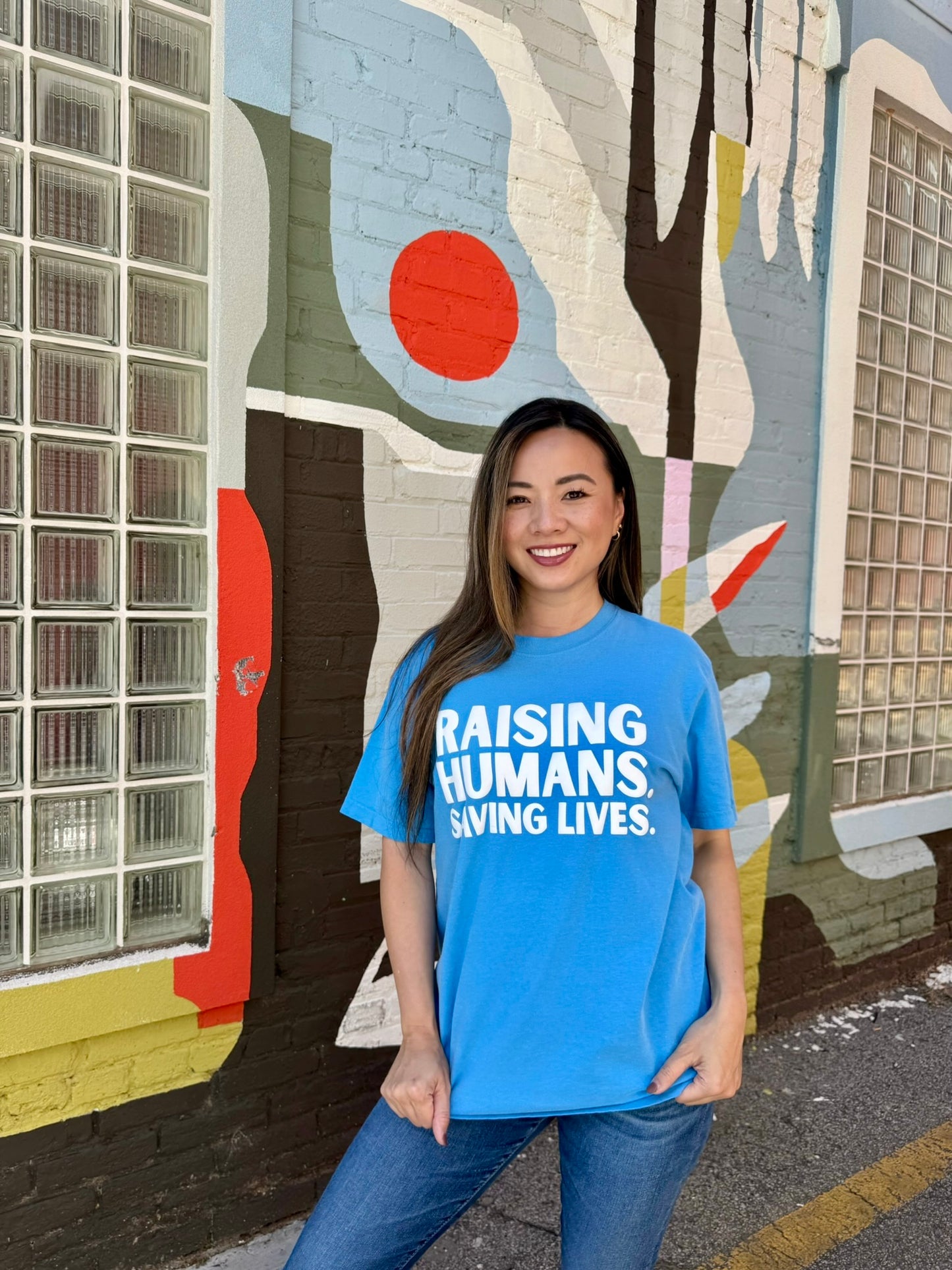 Woman wearing blue Raising Humans. Saving Lives. tee by Ladyspinedoc, standing in front of a mural with a bold, proud stance and smile.