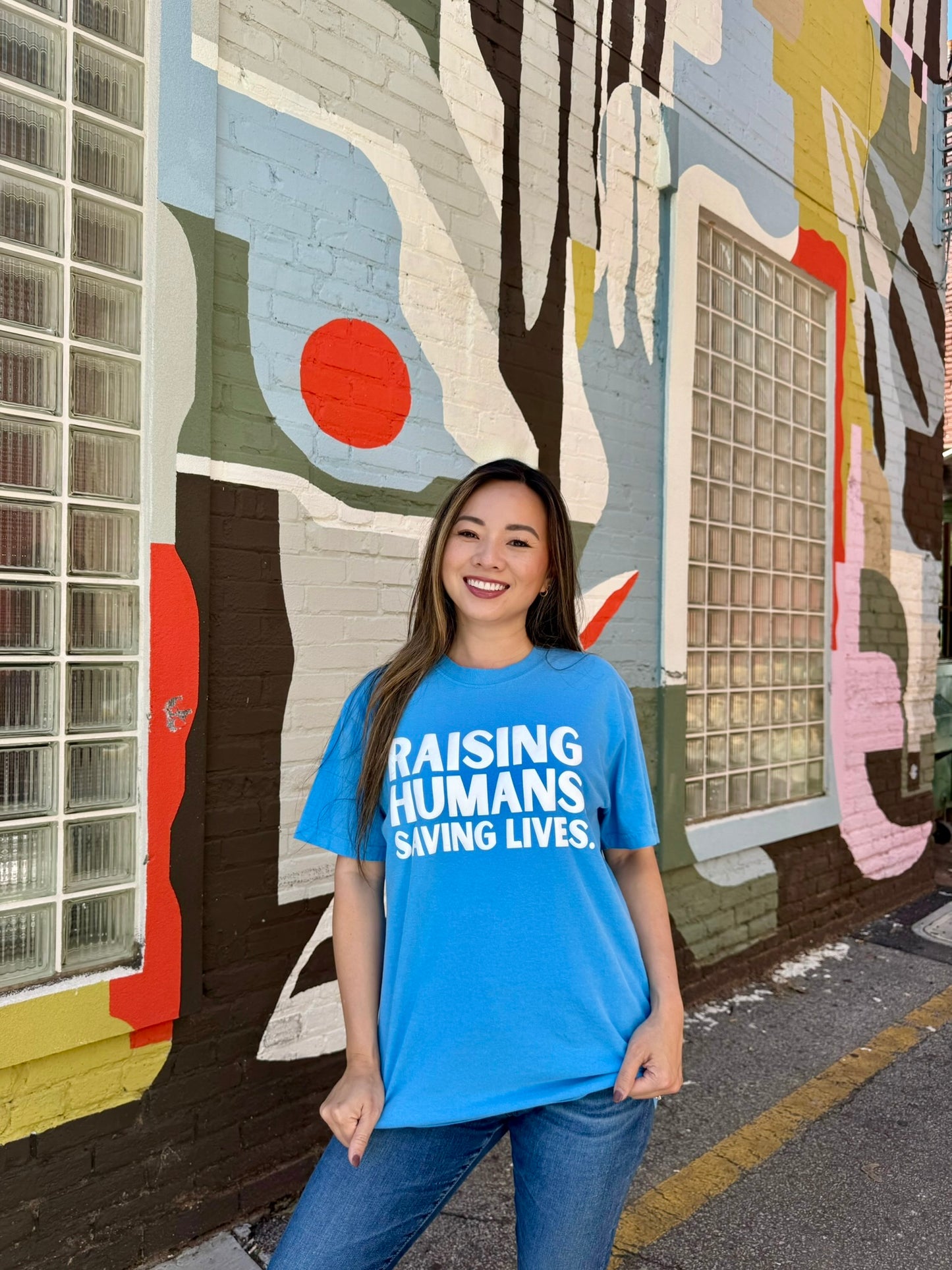 Woman wearing blue Raising Humans. Saving Lives. tee by Ladyspinedoc, standing in front of a mural and holding down the hem of the shirt.