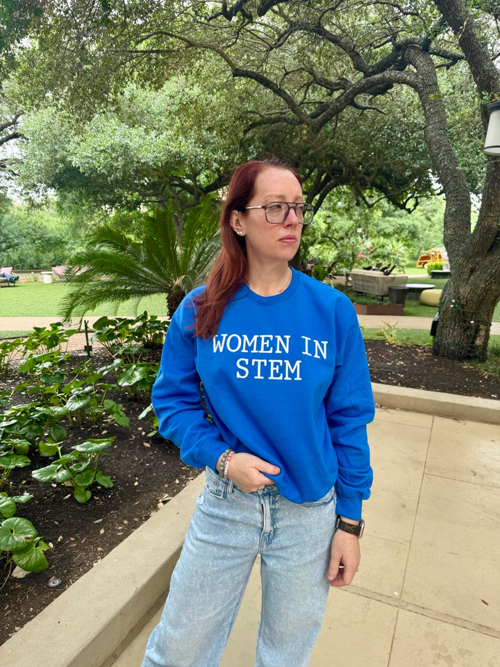 Ladyspinedoc stands outside in a garden setting wearing a blue "WOMEN IN STEM" sweatshirt, light-wash jeans, and glasses, surrounded by trees and greenery.