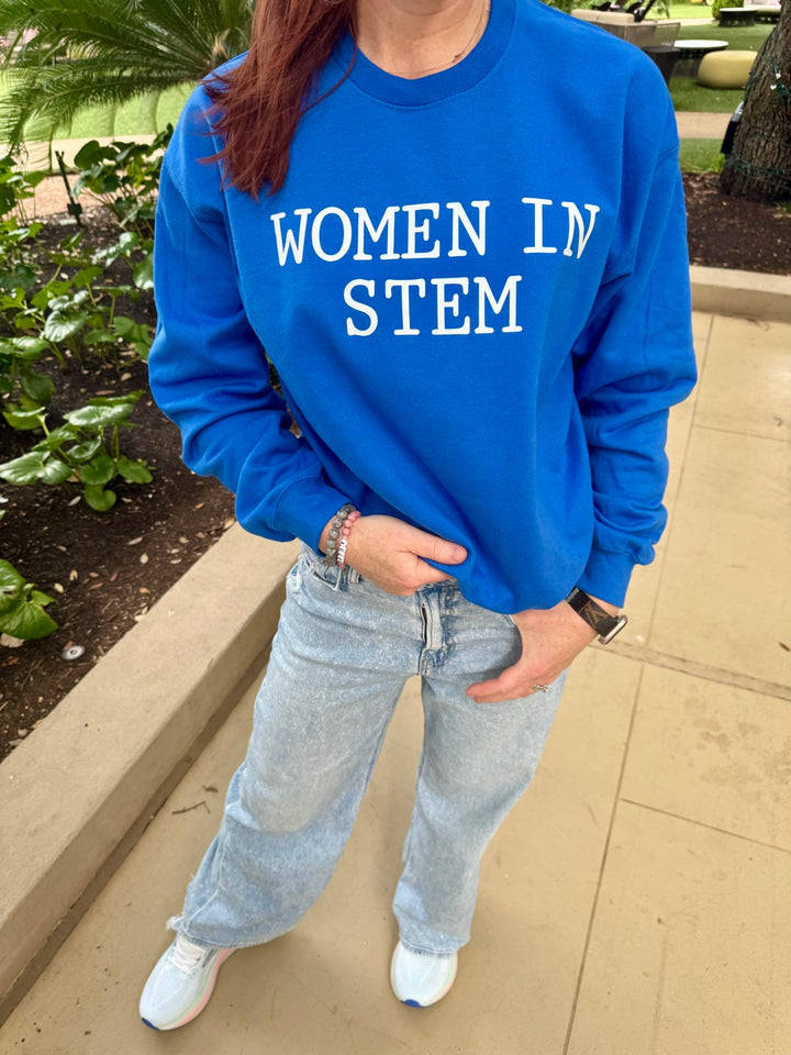 Close-up of Ladyspinedoc standing outdoors wearing a bright blue sweatshirt with bold white text that reads "WOMEN IN STEM," paired with light-wash jeans and white sneakers.