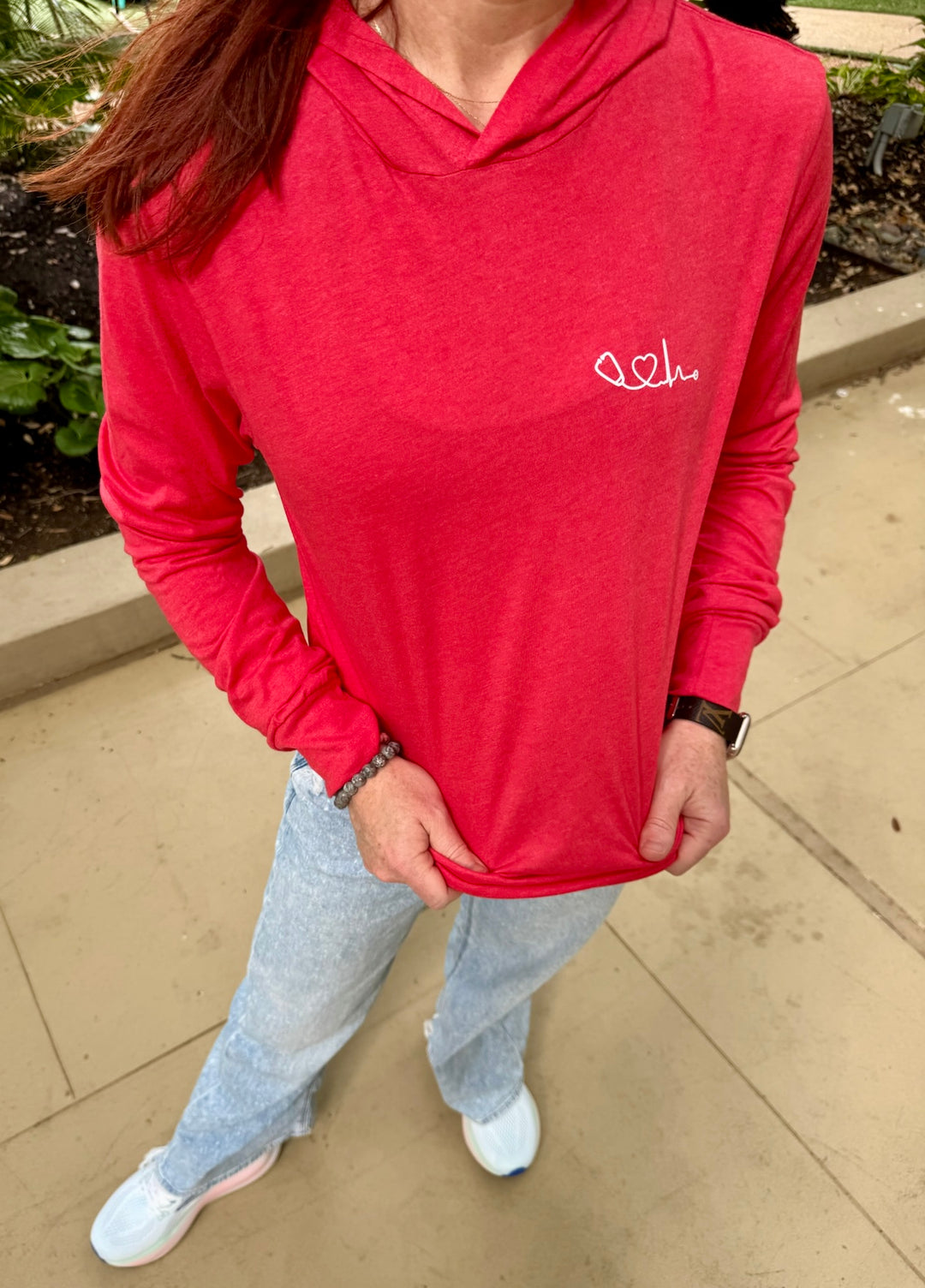 Dr. Betsy Grunch (Ladyspinedoc) wearing red long sleeve hoodie tee with Dr. Beachgem Heart of Medicine logo, front view outdoors.