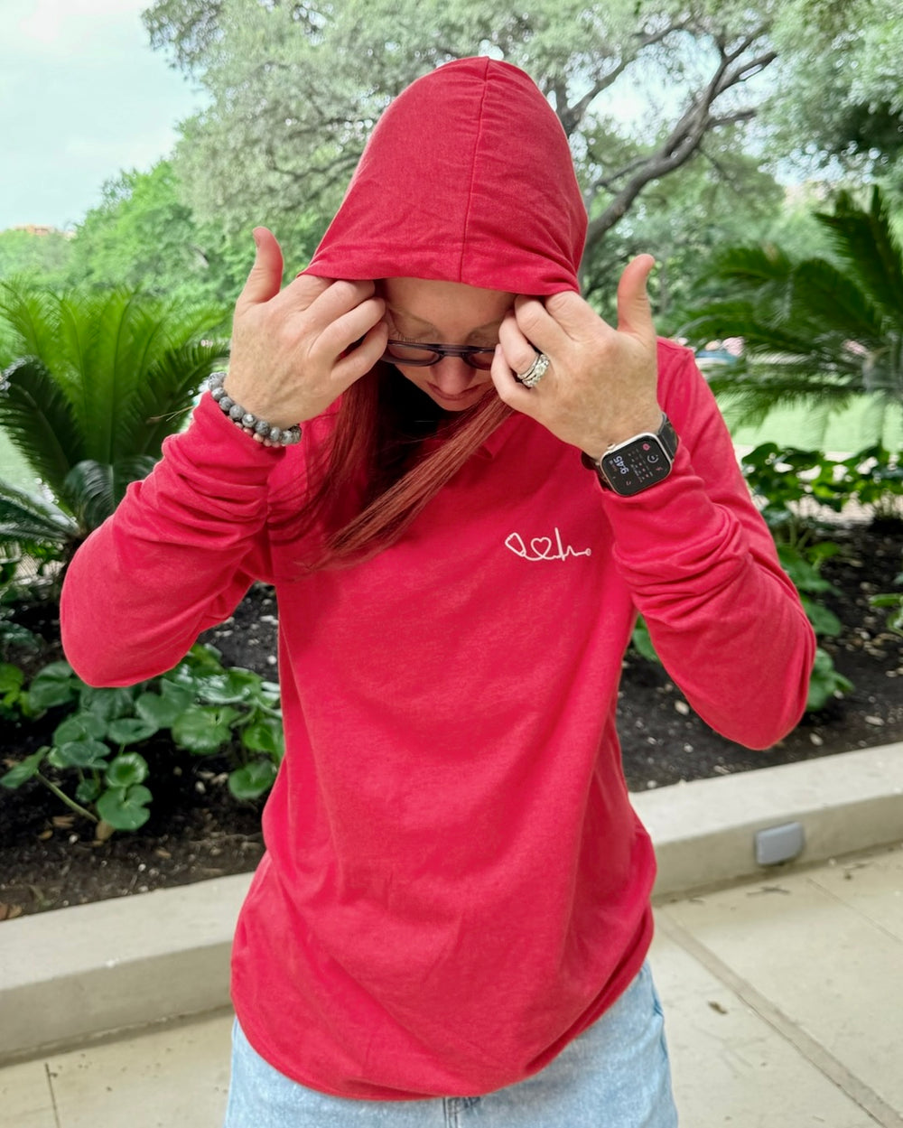 Dr. Betsy Grunch (Ladyspinedoc) in red Heart of Medicine tee with hood up, standing on patio with trees in background.