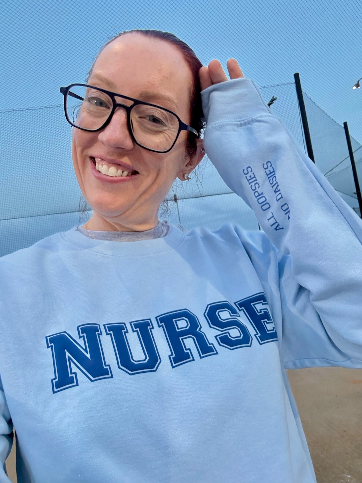 Smiling Ladyspinedoc (aka Dr. Betsy Grunch) wearing a light blue crewneck sweatshirt with “NURSE” printed on the front in bold varsity-style letters and “ALL OOPSIES NO DAISIES” on the left sleeve.