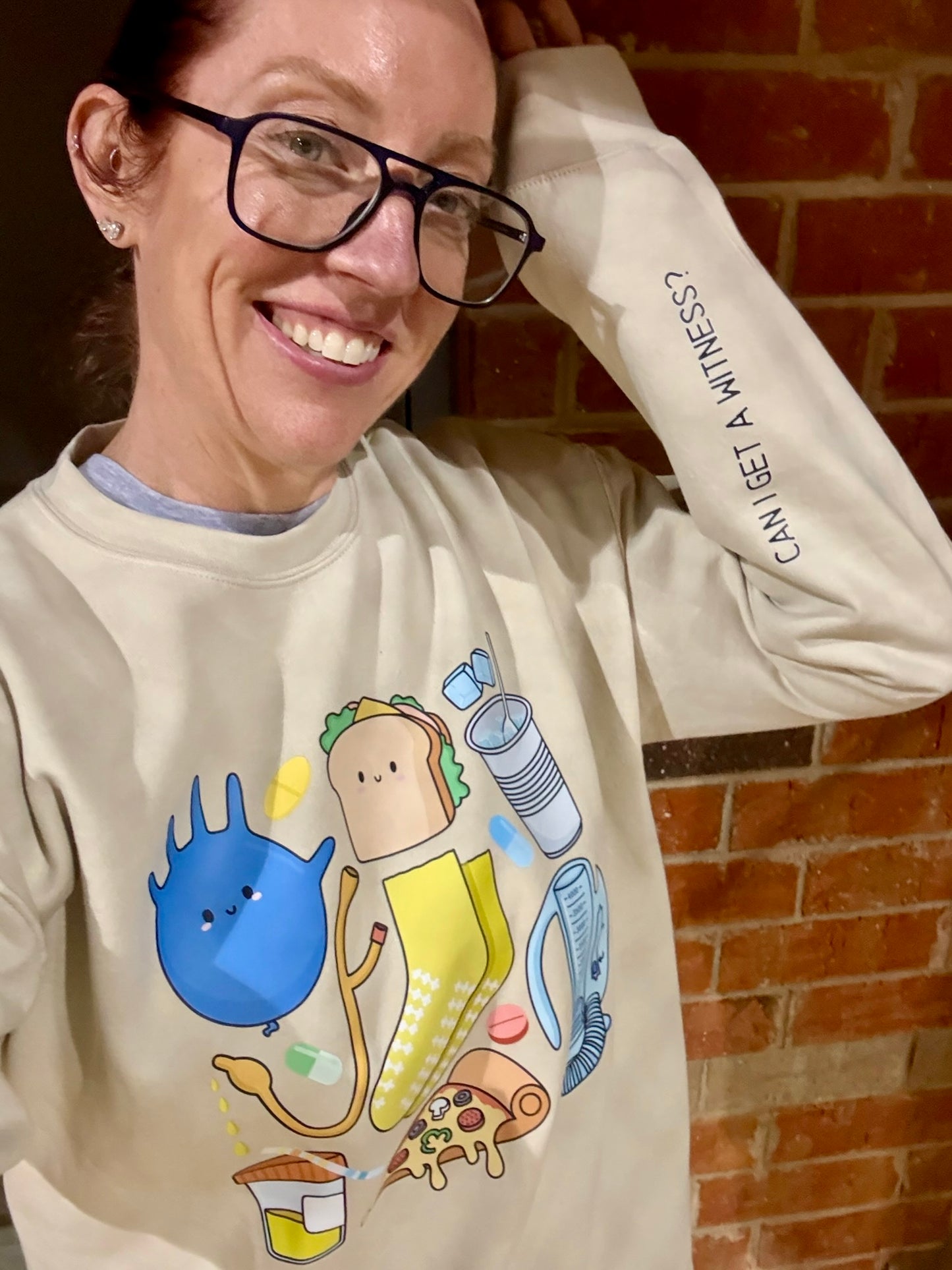 Ladyspinedoc (aka Dr. Betsy Grunch) wearing a sand-colored crewneck featuring colorful cartoon graphics of nurse essentials like a glove, meds, pizza, grippy socks, and more