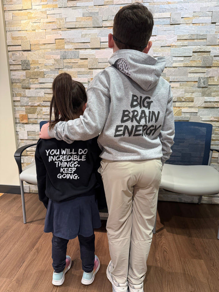 Girl youth model wearing the Incredible Youth Crewneck Sweatshirts, facing away and showing the back design with bold white brushstroke message: "YOU WILL DO INCREDIBLE THINGS. KEEP GOING." Male youth model wearing the youth Big Brain Energy Sweatshirt.