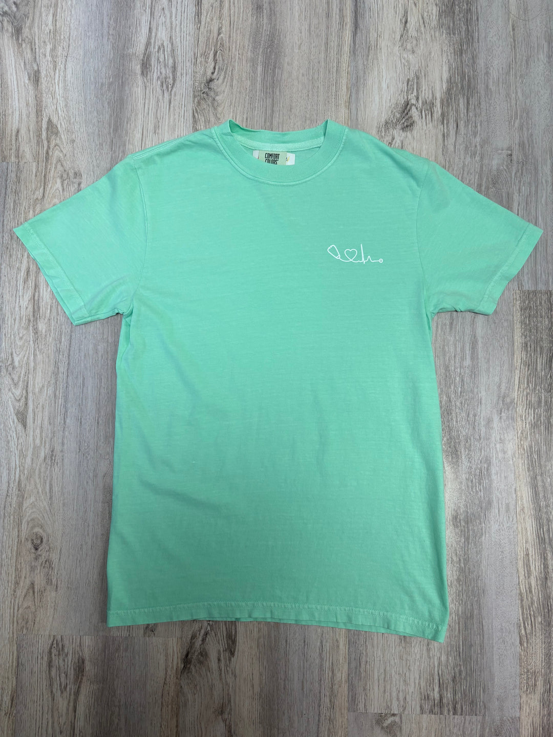 Flat lay of light green Heart of Medicine t-shirt featuring Dr. Beachgem’s stethoscope and heart waveform logo on the upper left chest.
