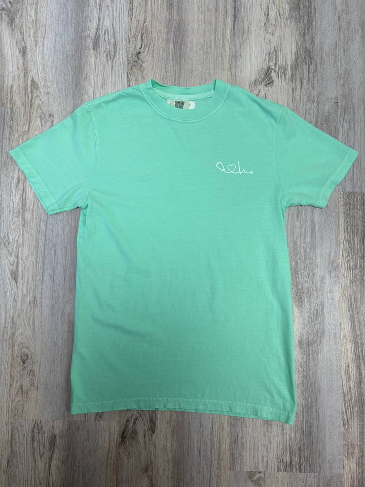Flat lay of light green Heart of Medicine t-shirt featuring Dr. Beachgem’s stethoscope and heart waveform logo on the upper left chest.