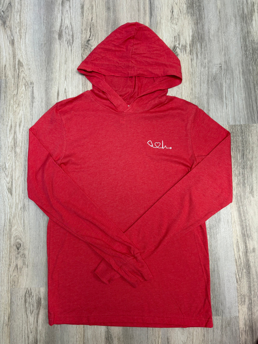 Flat lay of Heart of Medicine red long-sleeve hooded T-shirt with a white heart and stethoscope graphic on the chest, displayed on a wood floor — breathable medical-themed hoodie for nurses, doctors, and healthcare advocates.