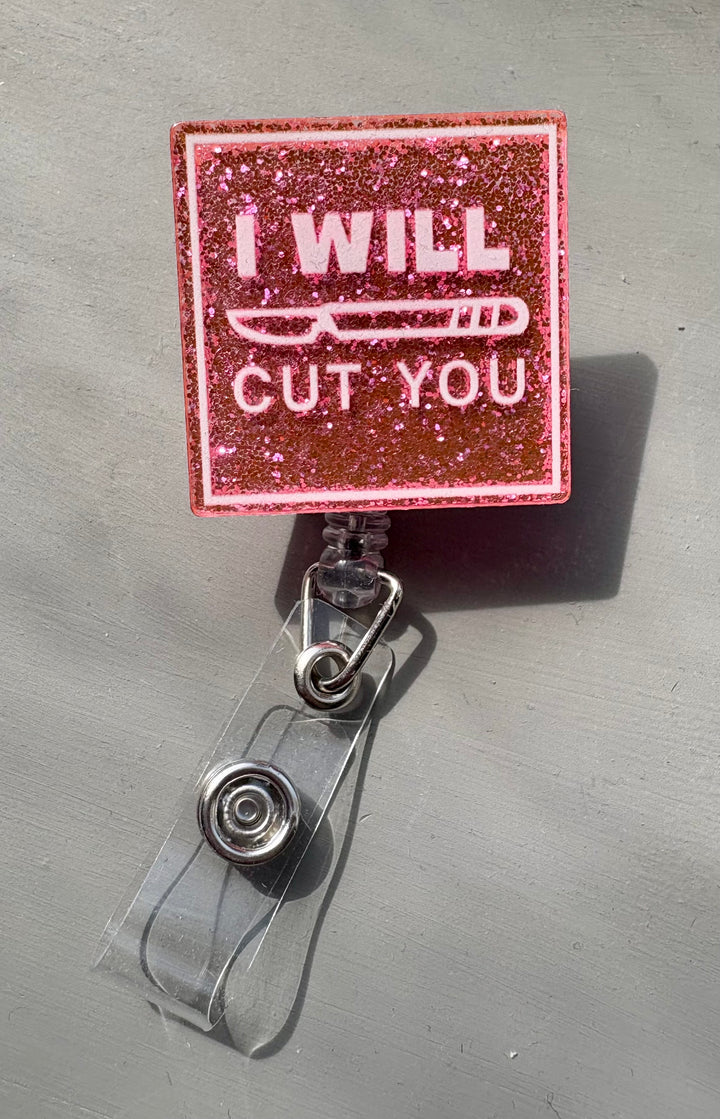 Pink glitter square badge reel with the phrase “I Will Cut You” printed in white, featuring a clear retractable clip for ID or name badge attachment.