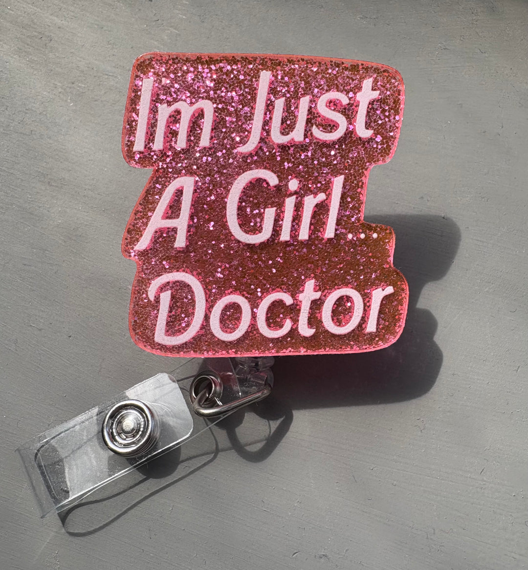 Retractable glitter pink badge reel with Barbie-style font that says “I'm Just A Girl Doctor.” Clip attachment shown below.