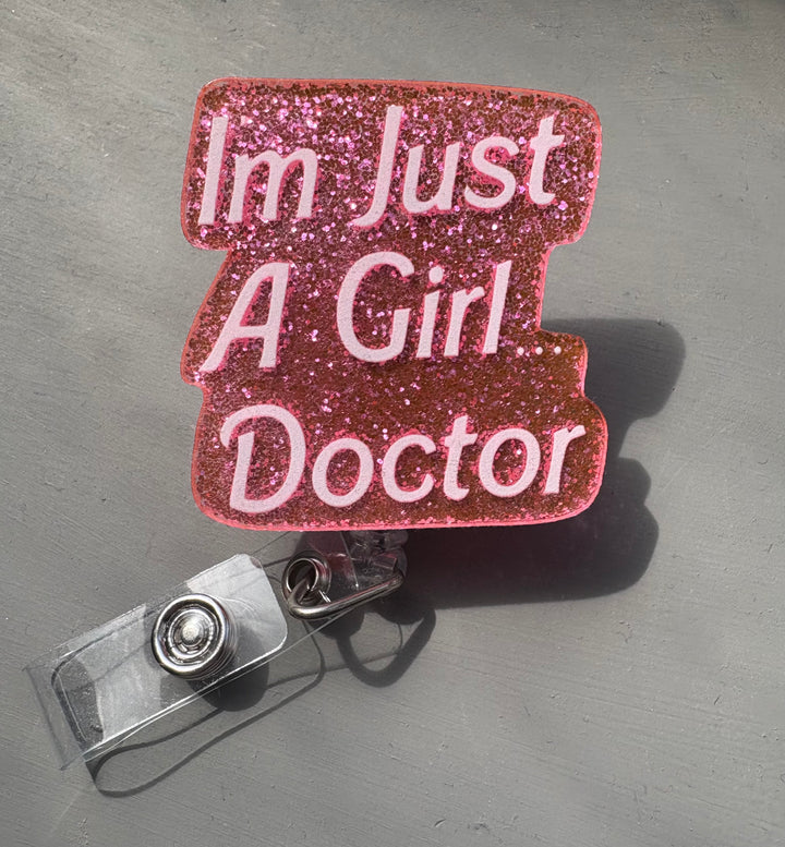 Retractable glitter pink badge reel with Barbie-style font that says “I'm Just A Girl Doctor.” Clip attachment shown below.