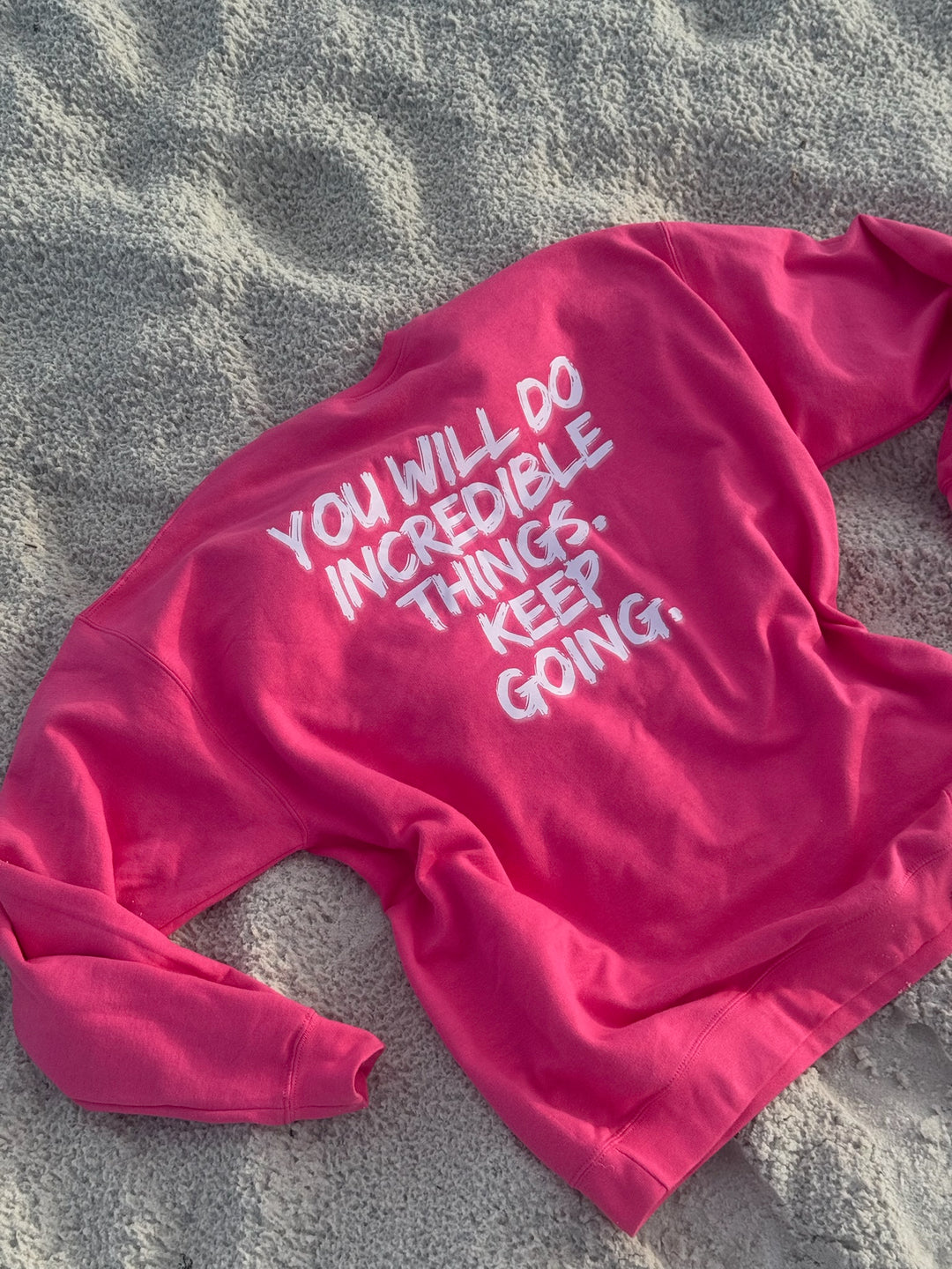 Pink “Incredible” sweatshirt by Ladyspinedoc laid flat on sandy beach, showing motivational quote on back in large white brushstroke font.