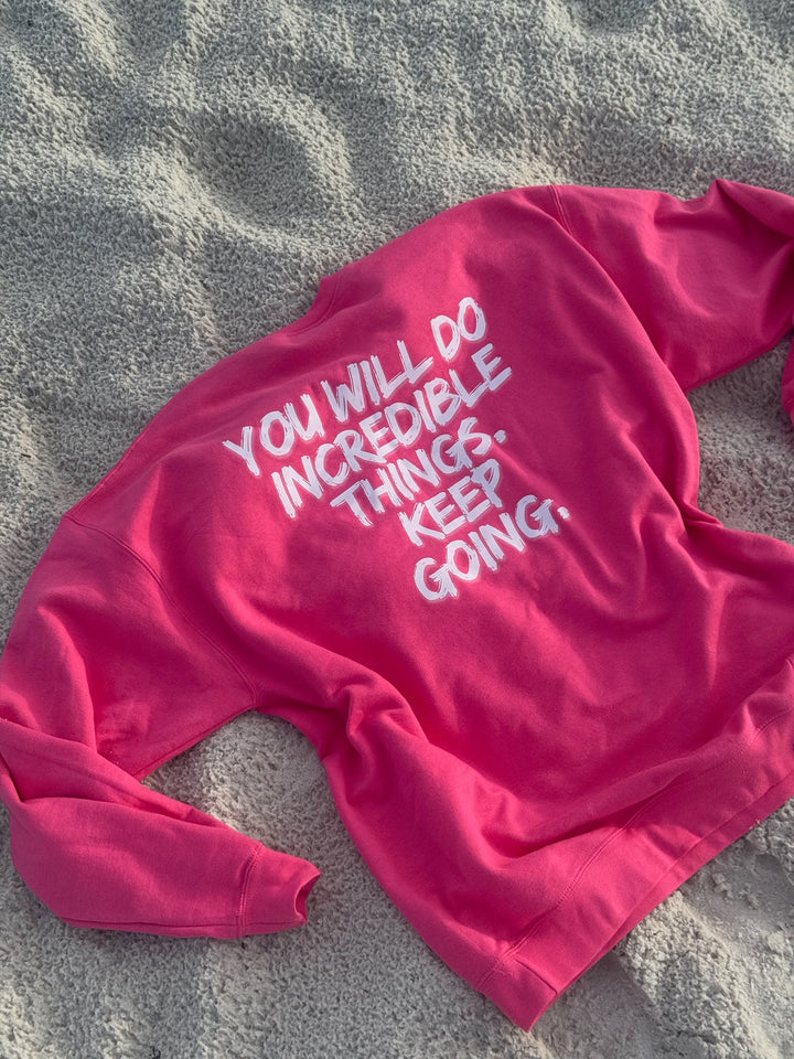 Pink “Incredible” sweatshirt by Ladyspinedoc laid flat on sandy beach, showing motivational quote on back in large white brushstroke font.