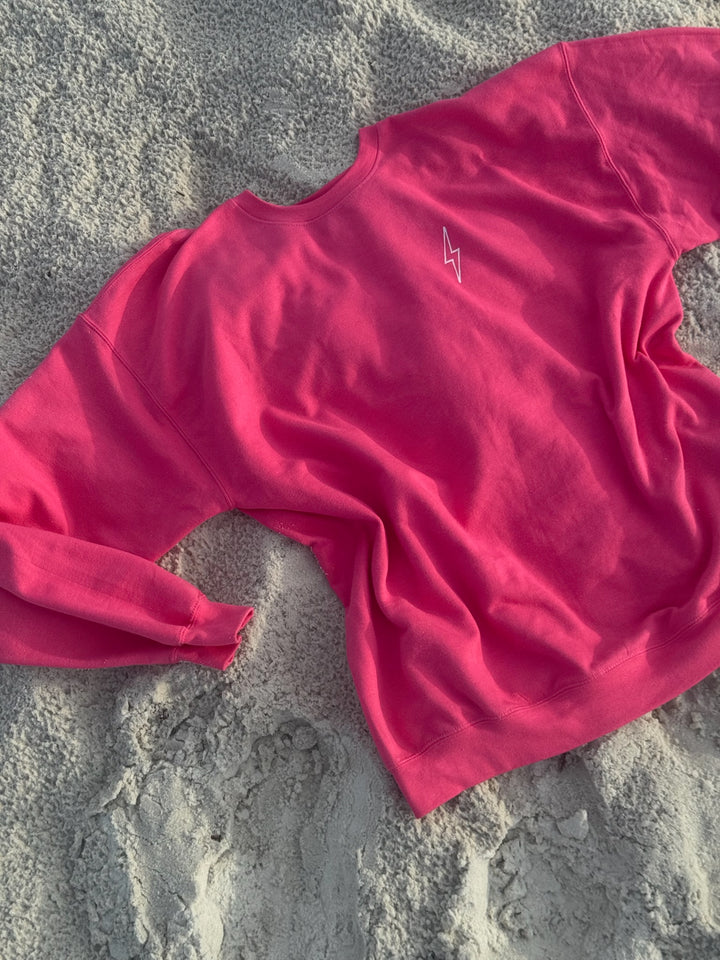 Front of Ladyspinedoc’s bright pink sweatshirt shown on the sand, featuring minimalist white lightning bolt design on upper left chest.