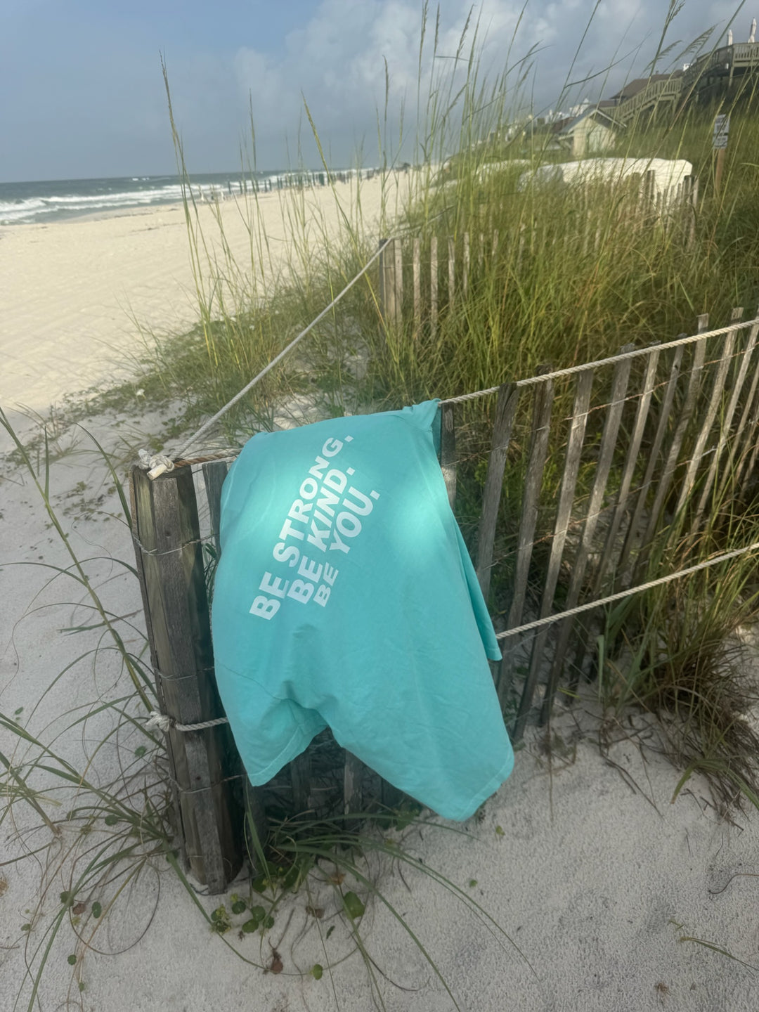 A "BE STRONG. BE KIND. BE YOU." T-shirt draped over a wooden beach fence, with sand dunes and sea oats in the background.