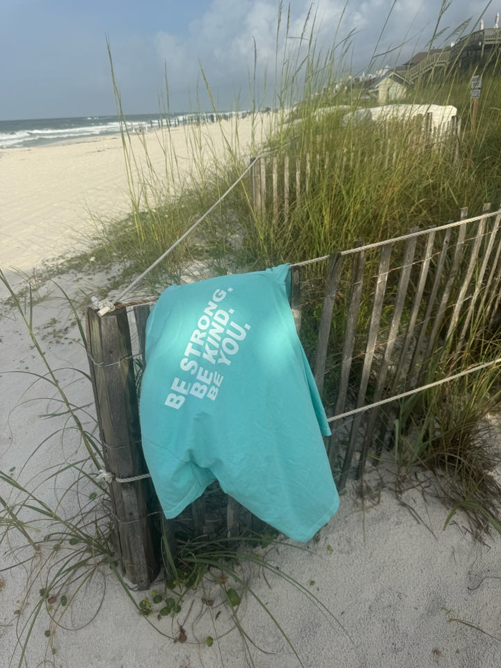 A "BE STRONG. BE KIND. BE YOU." T-shirt draped over a wooden beach fence, with sand dunes and sea oats in the background.