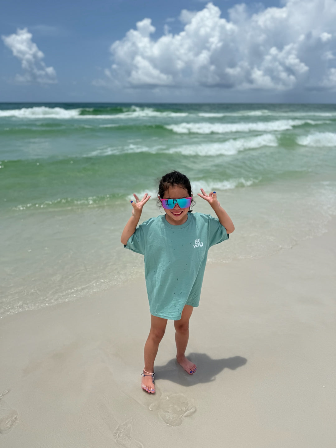 A smiling young girl in oversized sunglasses poses with peace signs on a sandy beach, wearing a light blue "BE YOU" tee with the ocean and clouds behind her.