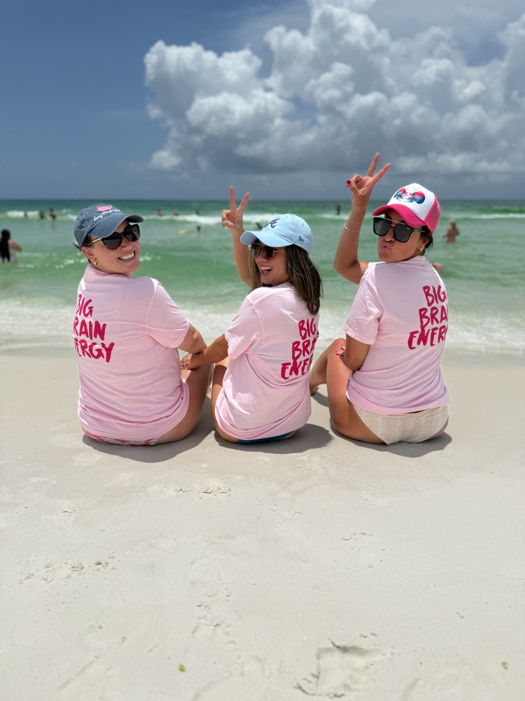 Three women sitting on the beach facing the ocean, all wearing matching “Big Brain Energy” pink T-shirts with bright pink text on the back. They’re smiling and flashing peace signs—fun, empowering beachwear for strong women in STEM and healthcare.