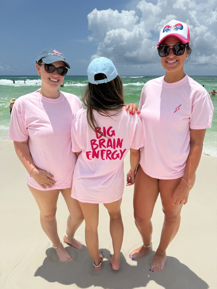 Three women standing on the beach wearing matching light pink “Big Brain Energy” T-shirts. Two face forward showing the front lightning bolt logo, while the center person shows the bold pink “Big Brain Energy” text on the back. Perfect for healthcare professionals and empowered women.
