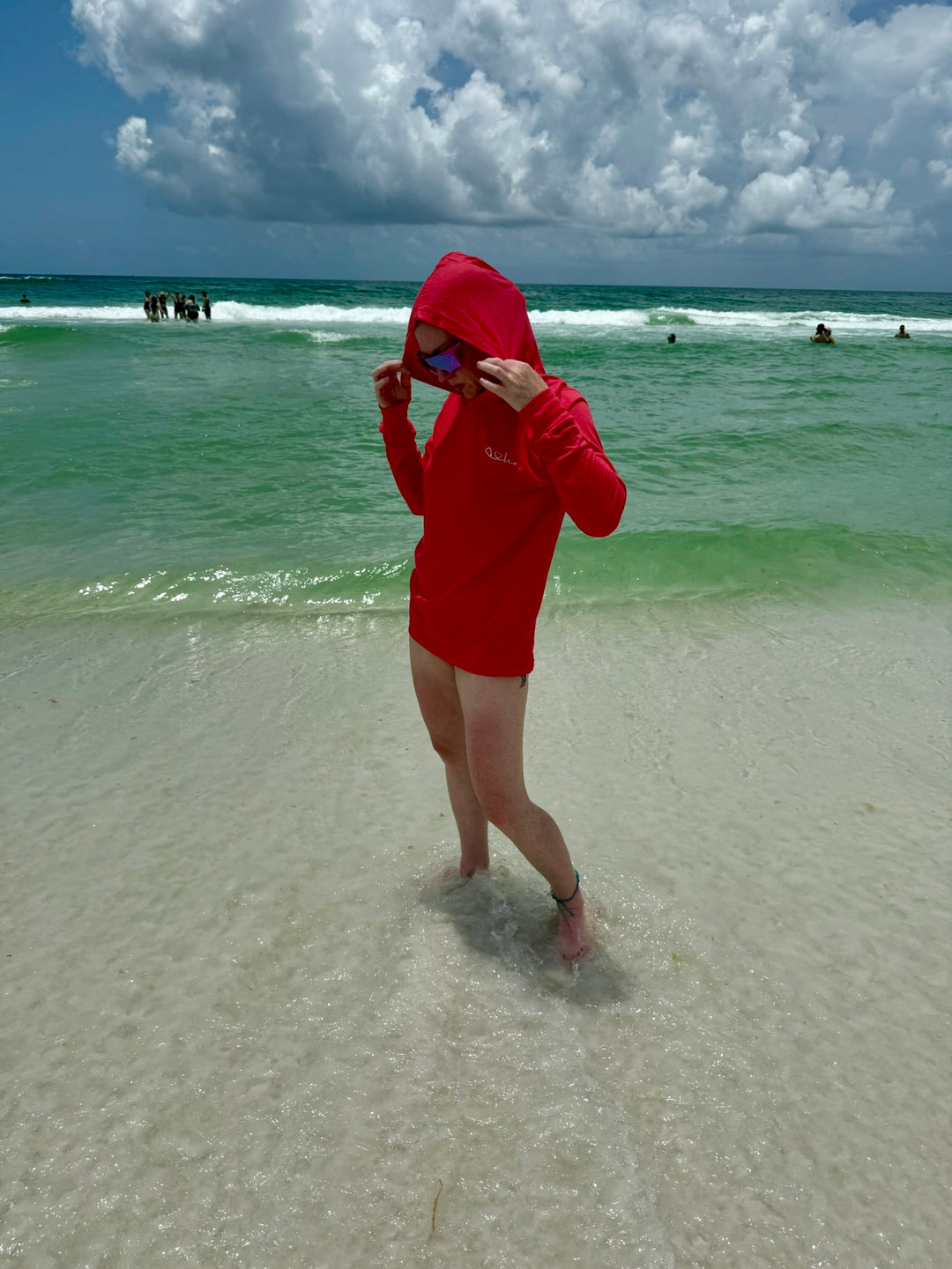 Ladyspinedoc standing ankle-deep in ocean water wearing Heart of Medicine  lightweight red hooded long-sleeve shirt with a white stethoscope heart design on the chest — beach-ready sun protection for healthcare workers and medical professionals.