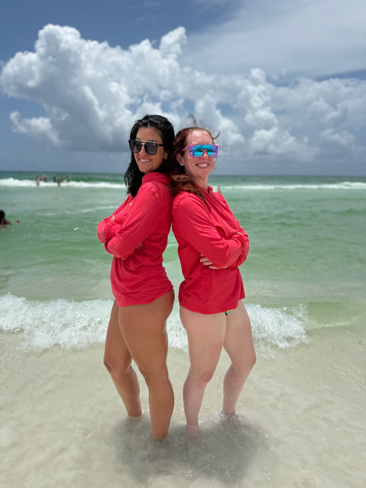 Two smiling women standing back-to-back at the beach wearing matching red  Heart of Medicine lightweight hoodies with stethoscope heart designs — perfect medical apparel for sunny days and strong women in medicine.