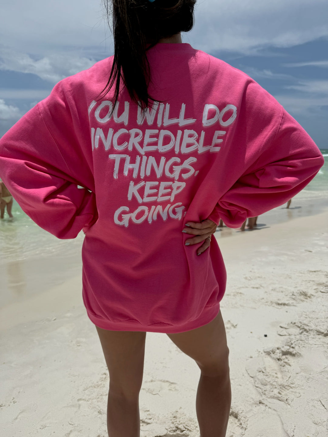 Back view of woman wearing bright pink Ladyspinedoc sweatshirt on the beach, featuring bold white text that reads “YOU WILL DO INCREDIBLE THINGS. KEEP GOING.”