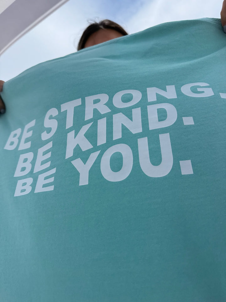 A close-up of a light blue T-shirt held up to display the bold white text: "BE STRONG. BE KIND. BE YOU." on the back.