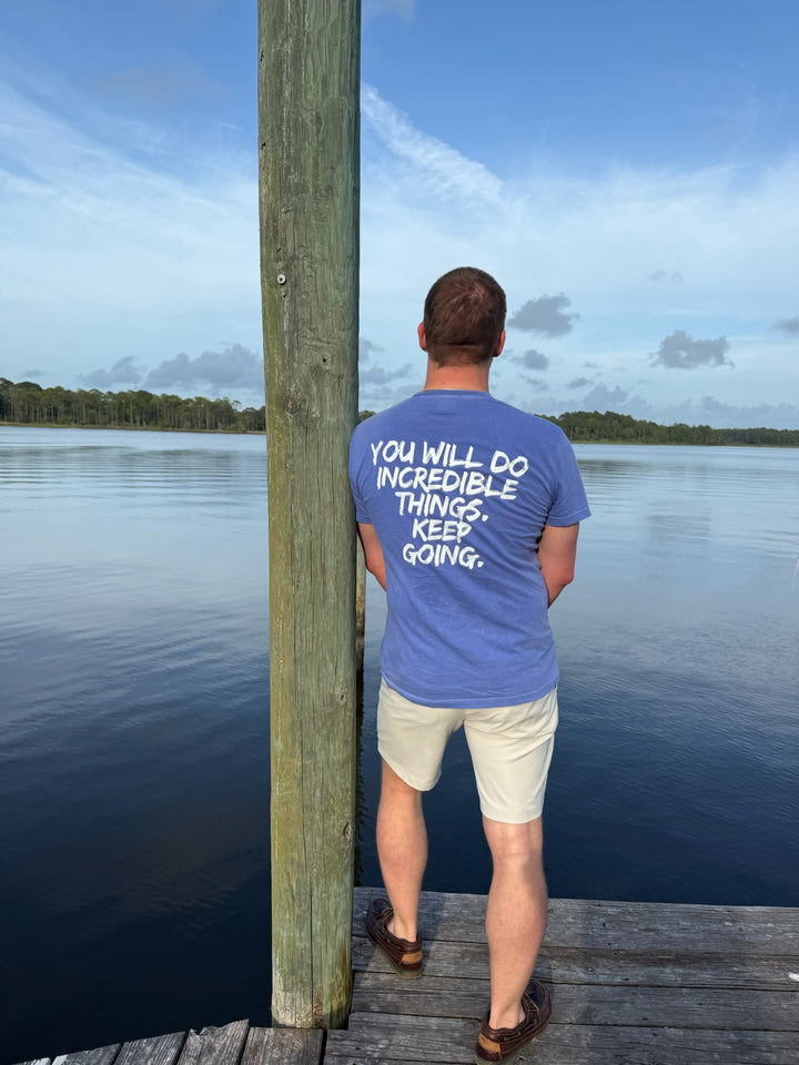 Male at waterfront showing the back of purple tee with bold white text: “YOU WILL DO INCREDIBLE THINGS. KEEP GOING.”