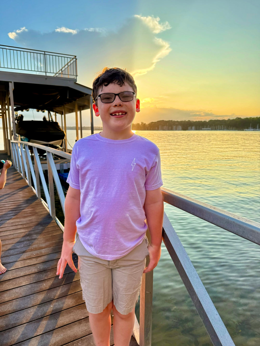 Kid at the lake at sunset wearing the violet Incredible Youth Tee, showing the front with Ladyspinedoc signature lightning bolt displayed on upper left chest.
