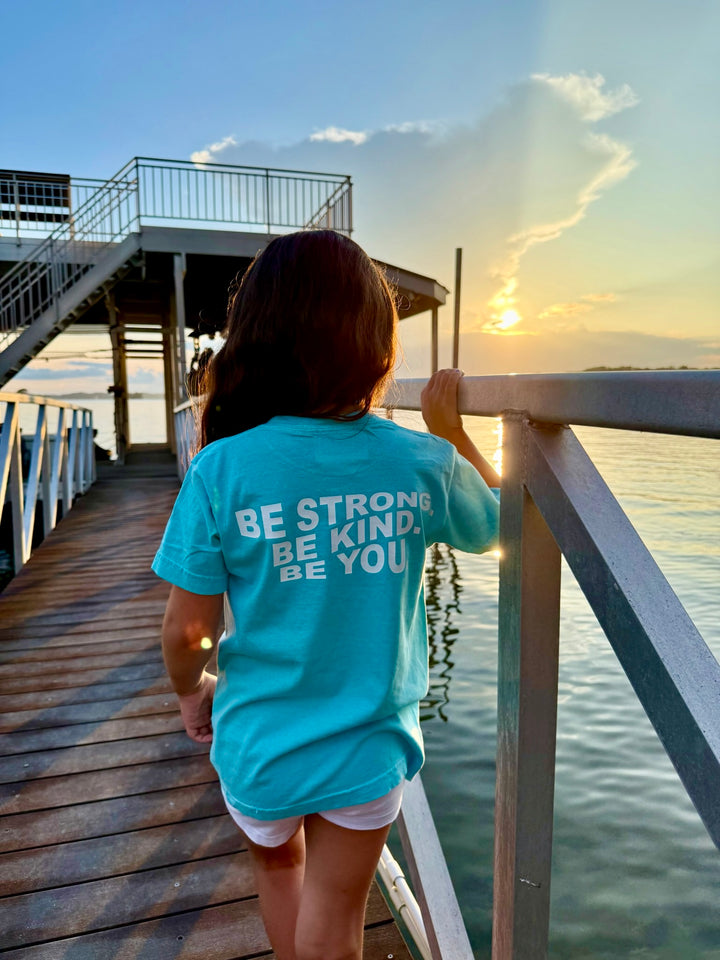 Young girl in a light blue Ladyspinedoc Be You Youth Tee walking on a dock at sunset, showing the bold back design that reads “BE STRONG. BE KIND. BE YOU.”