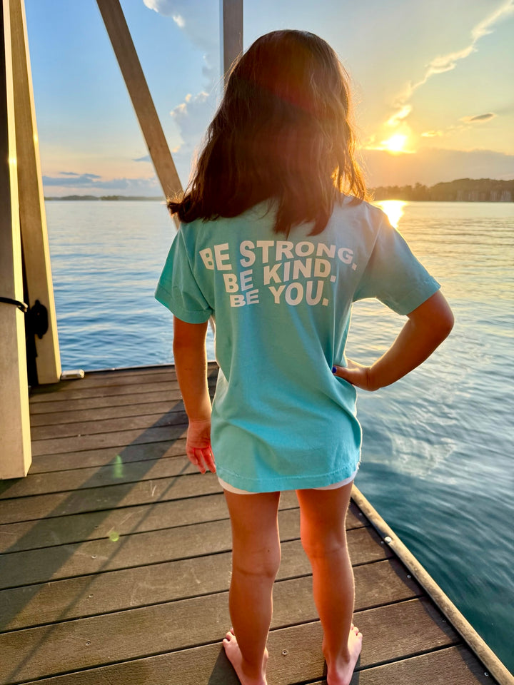 Child standing confidently on a dock in the Be You Youth Tee, with the motivational phrase “BE STRONG. BE KIND. BE YOU.” clearly visible on the back of the shirt.