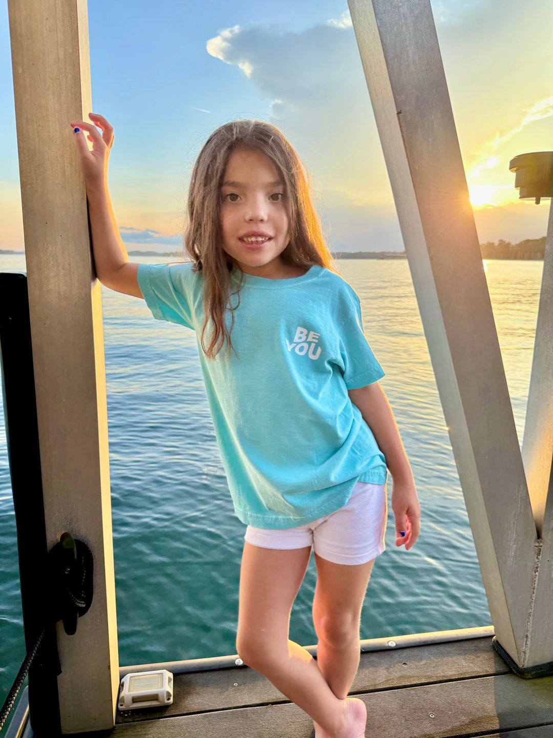 Smiling girl posing in front of a lake at sunset, wearing the light blue Be You Youth Tee by Ladyspinedoc, featuring “BE YOU” on the upper left chest.