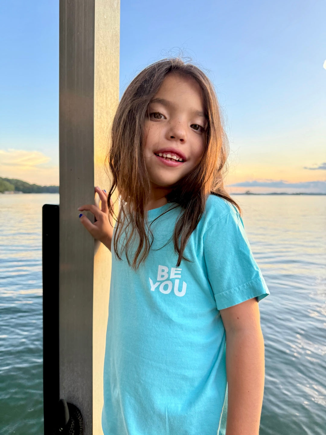 Girl leaning on a dock post at sunset, wearing the light blue Be You Youth Tee by Ladyspinedoc, with “BE YOU” printed on the front chest.