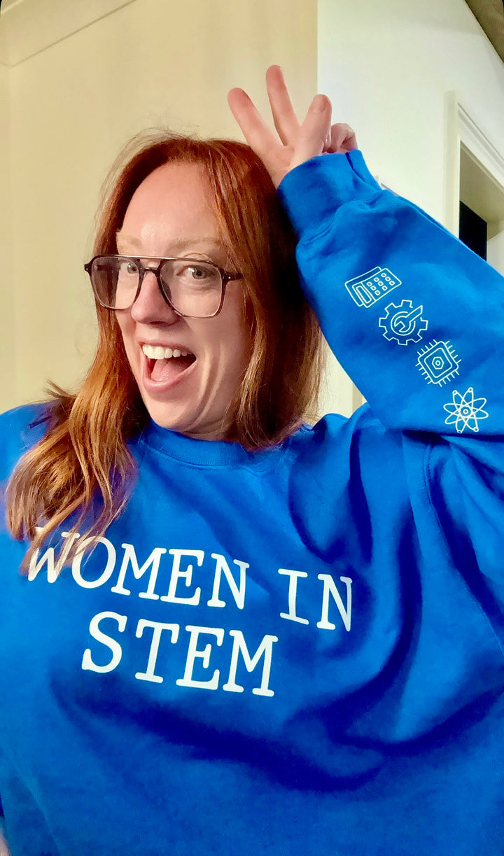 Ladyspinedoc wearing a bright blue sweatshirt with bold white text that reads "WOMEN IN STEM," and showing off the left sleeve with STEM icons.