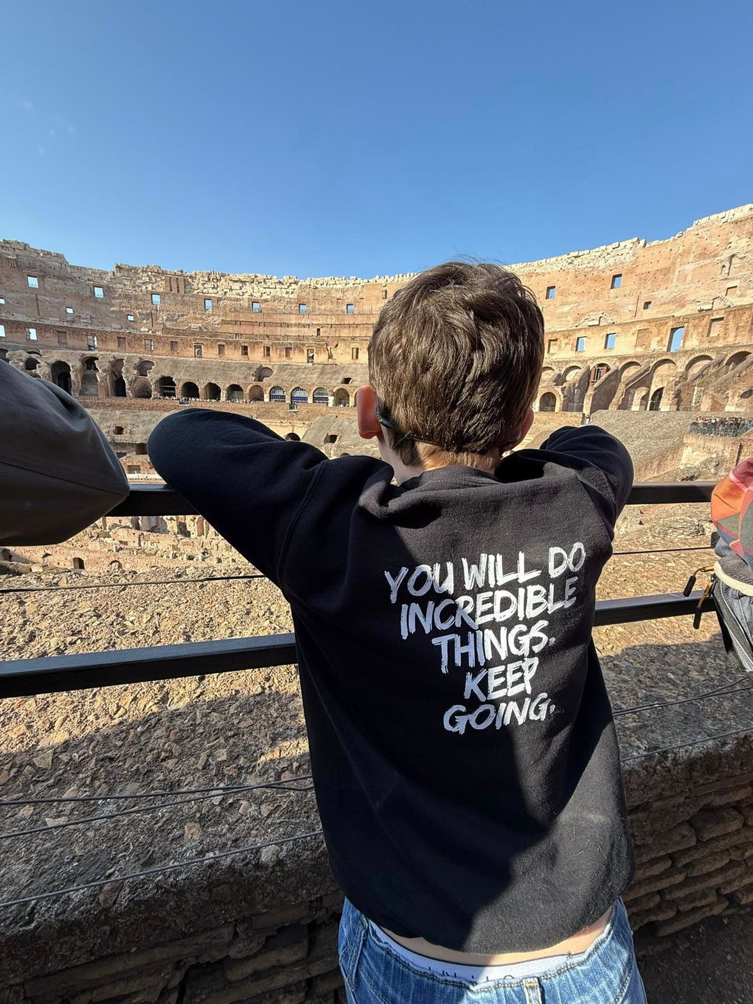 Back of Male Youth staring at The Colosseum in Rome and wearing the Kids Incredible Sweatshirt with "YOU WILL DO INCREDIBLE THINGS. KEEP GOING." on the back.