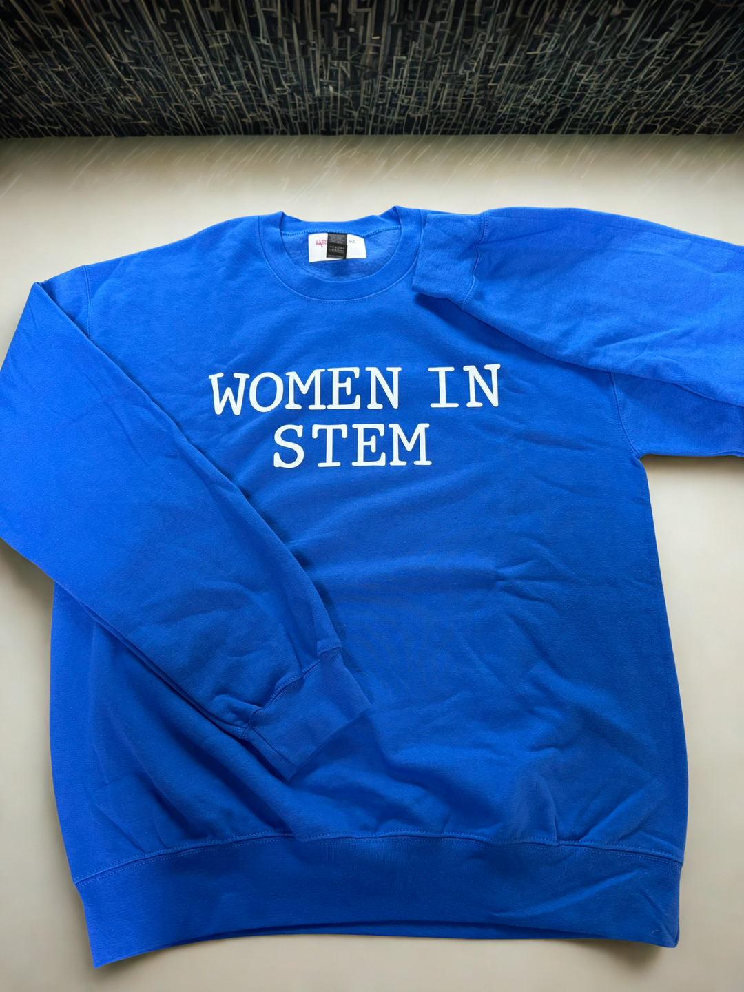 Royal blue crewneck sweatshirt with bold white text reading “Women in STEM,” laid flat on a counter