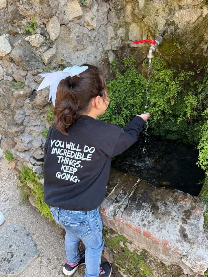 Back of young girl at a water spout in Rome, Italy wearing the Kids Incredible Sweatshirt with "YOU WILL DO INCREDIBLE THINGS. KEEP GOING."  motto.