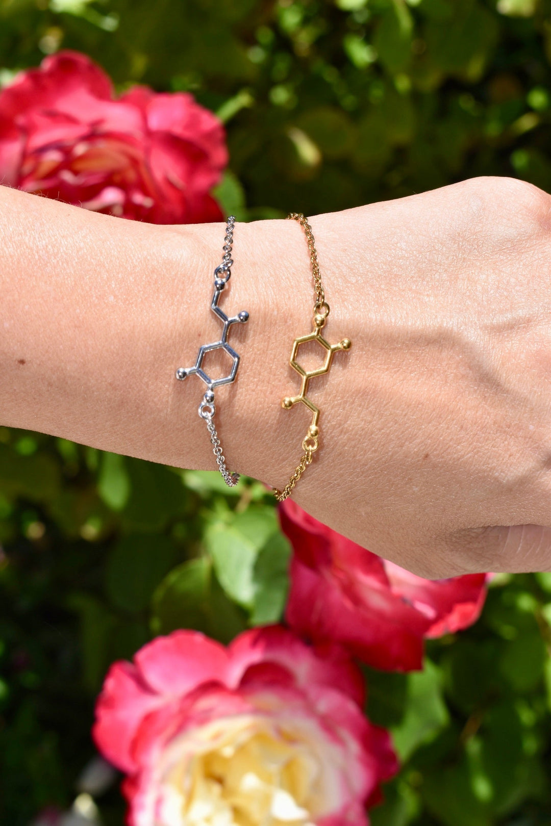 Silver and gold Noradrenaline molecule bracelets on a wrist with pink and yellow flowers in the background