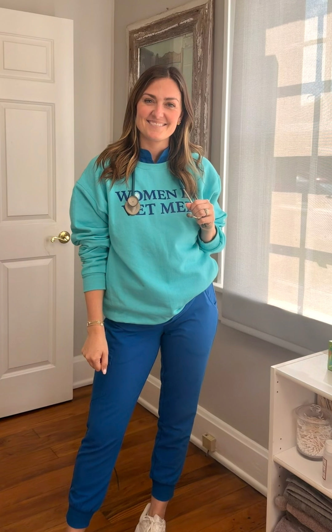 Dr Molly Brinkmann wearing a teal sweatshirt with women in vet med text in a room with a door and window.