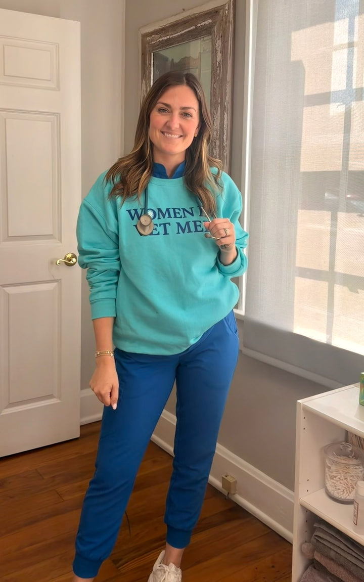 Dr Molly Brinkmann wearing a teal sweatshirt with women in vet med text in a room with a door and window.