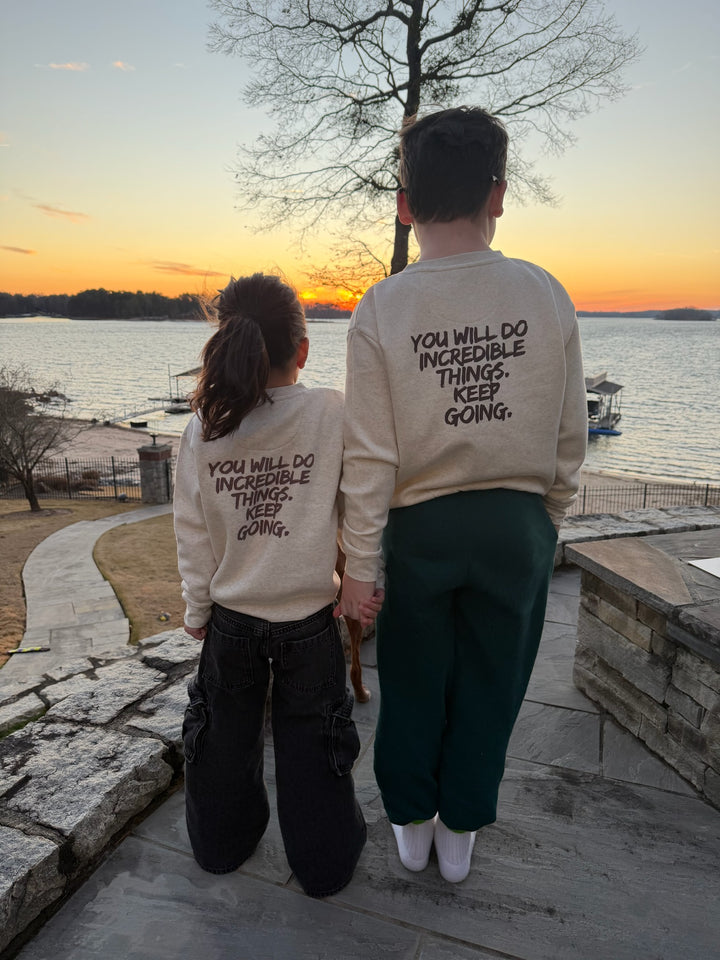 Two youths wearing matching Incredible Sweatshirts - Kids Natural Heather Shirt with Motivational "YOU WILL DO INCREDIBLE THINGS. KEEP GOING." motto on the back, standing by a lake at sunset.