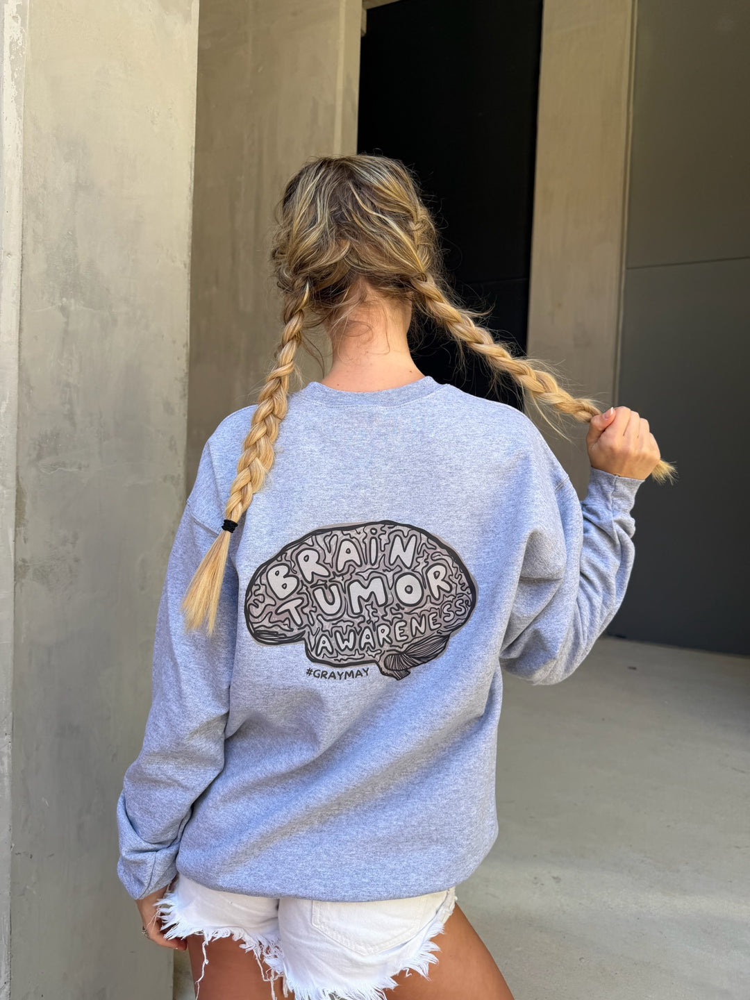 Person wearing a gray sweatshirt with a brain tumor awareness design, standing against a neutral wall.