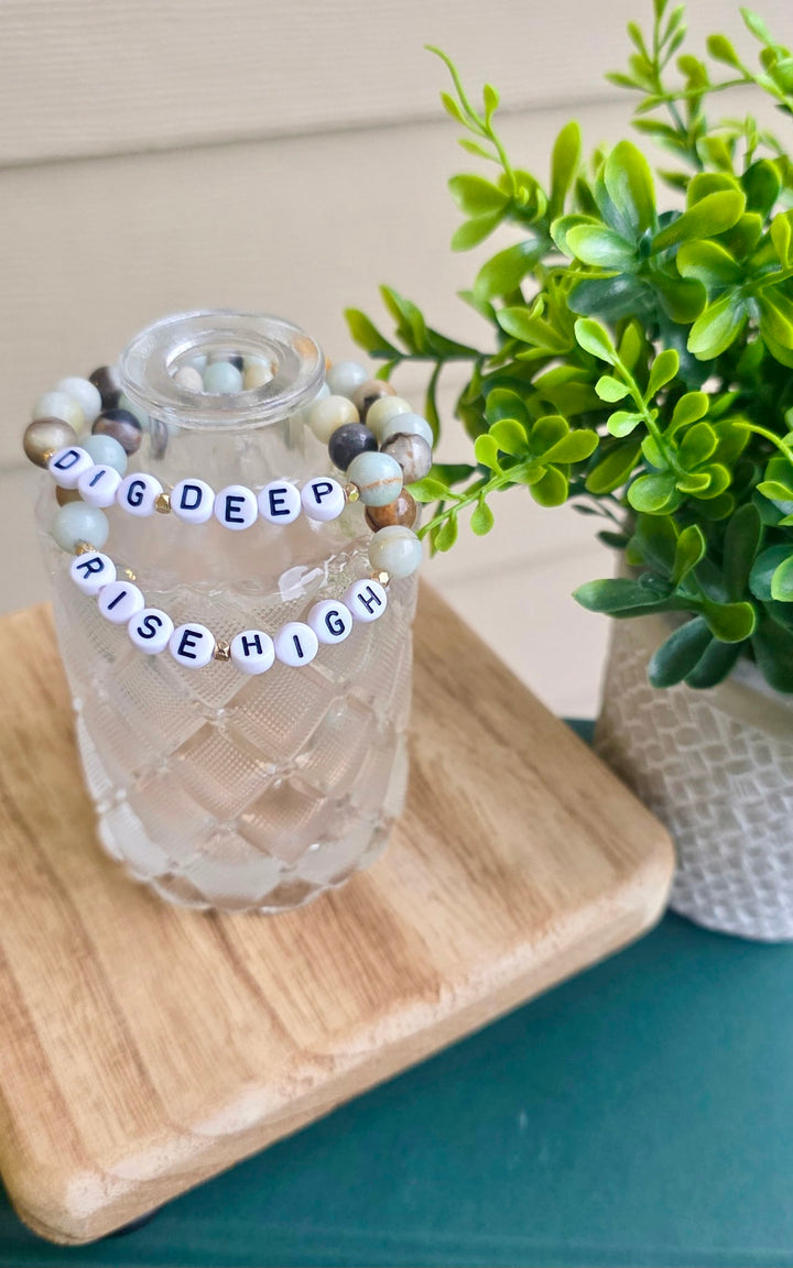 Glass vase with Dr. Potter's "Dig Deep Rise High" beaded bracelets  on a wooden coaster with plant in background