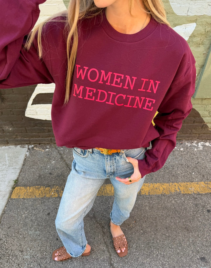 Person wearing a maroon sweatshirt with pink 'WOMEN IN MEDICINE' text on a street.